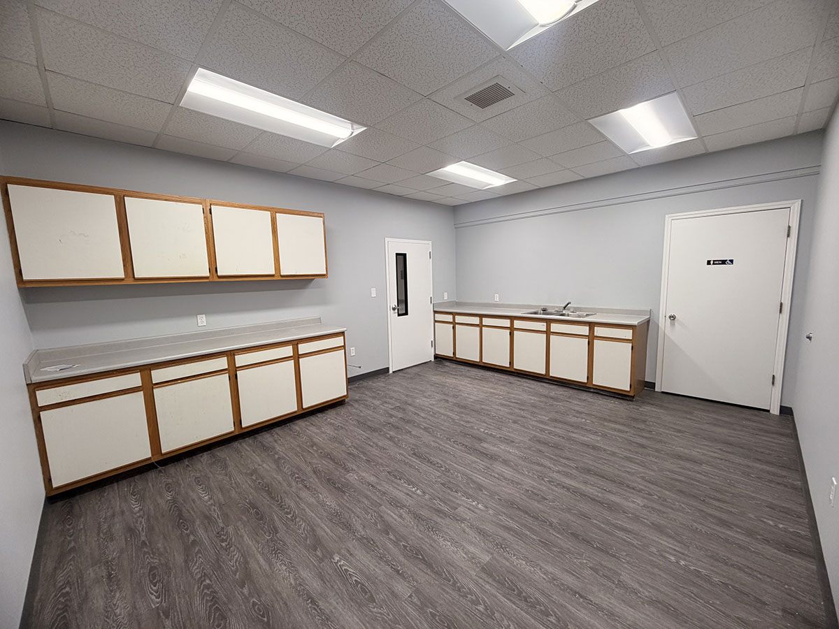 Large break room at 1310 Starr Drive with two long runs of cabinets and counter, a double sink, grey vinyl plank flooring, and a restroom door off the far corner