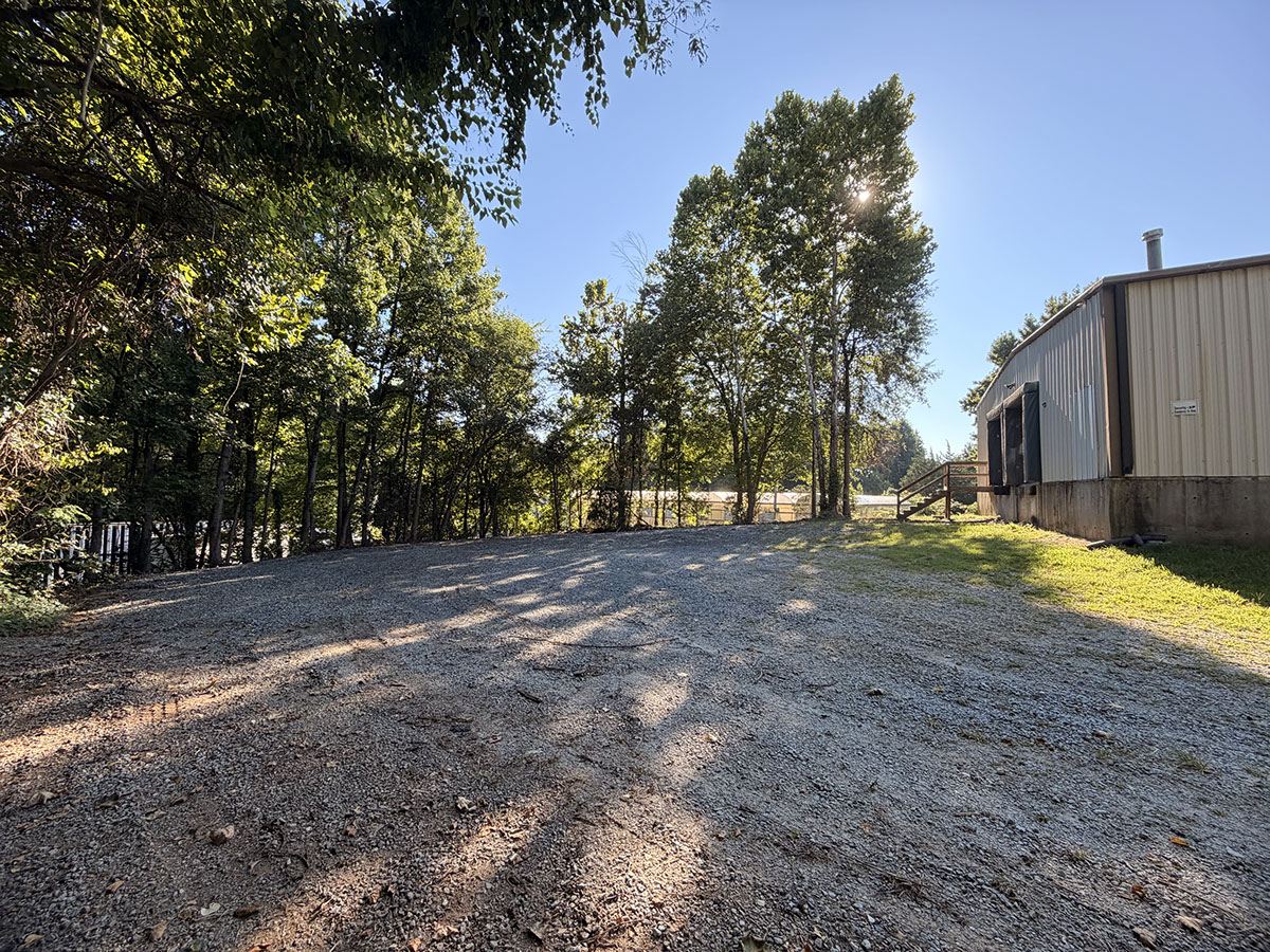 The gravel dock court alongside 1310 Starr Drive, an open graded area running the length of the building with a dock door and steps at the right and a tree line beyond
