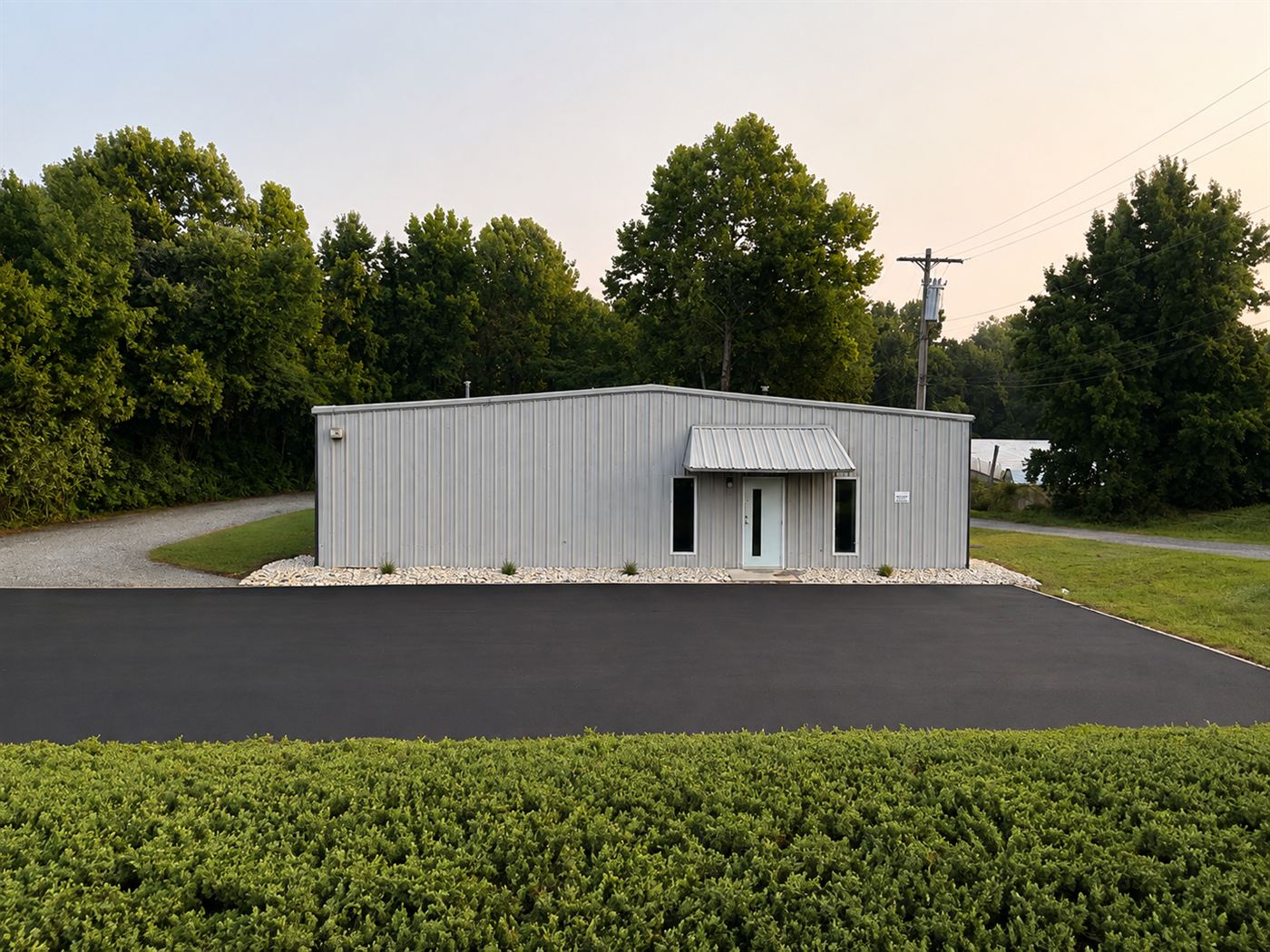 Front elevation of 1310 Starr Drive: a light grey metal building with a standing-seam canopy over the glazed entry, flanking windows, a white stone bed along the base, and fresh asphalt in front