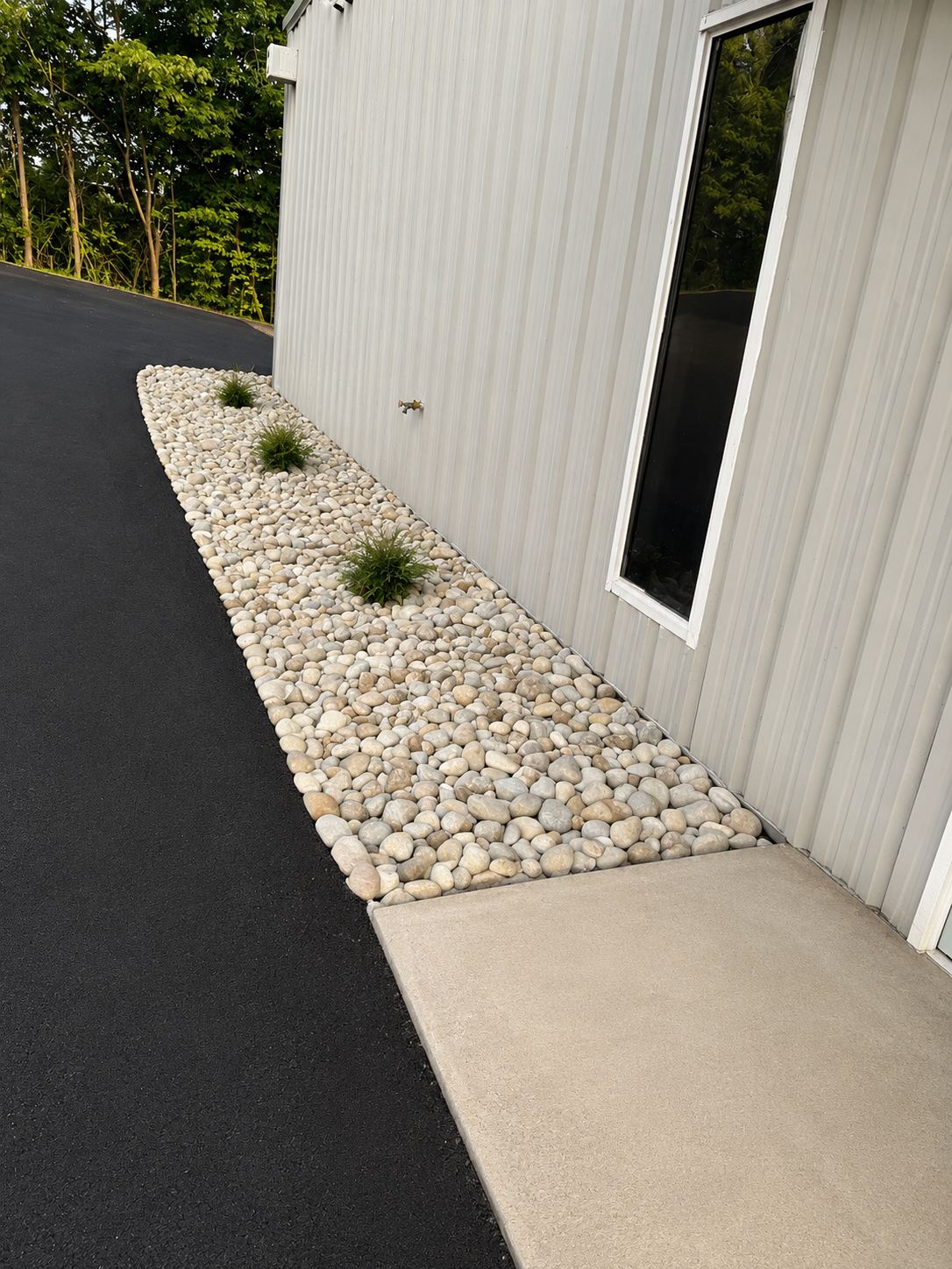 Landscaped white stone bed with ornamental grasses running along the base of the metal wall at 1310 Starr Drive, edging fresh asphalt beside a concrete pad