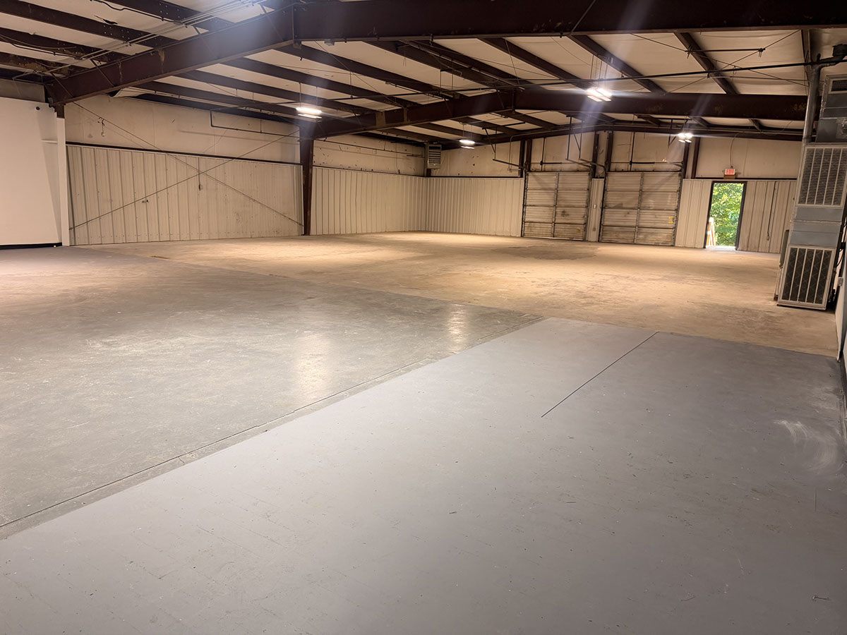 Interior of the warehouse at 1310 Starr Drive: an open concrete floor, light metal walls, exposed steel roof framing, overhead lighting, and a personnel door open to daylight at the far end