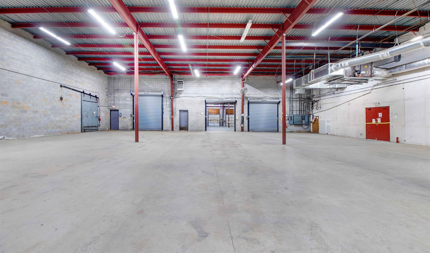 Warehouse interior at 1904 New Hope Church Road with a broad concrete floor, painted block walls, red steel columns and roof framing, two roll-up doors, and a passage through to the dock area