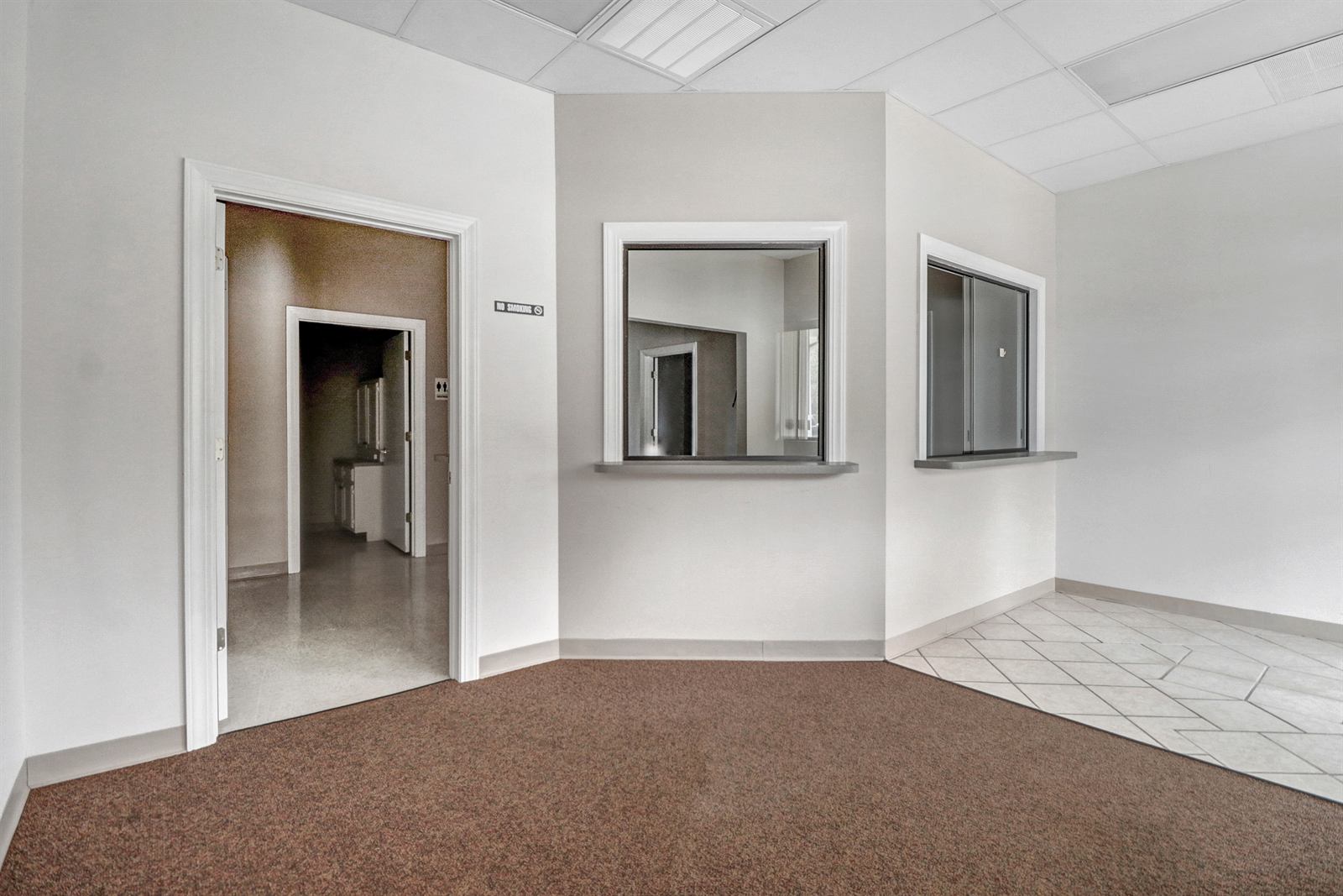 Reception area of the available suite at 1990 Allen Road, with two glazed transaction windows set into a partition wall, carpeted waiting area, tiled entry, drop ceiling, and a doorway through to the restroom