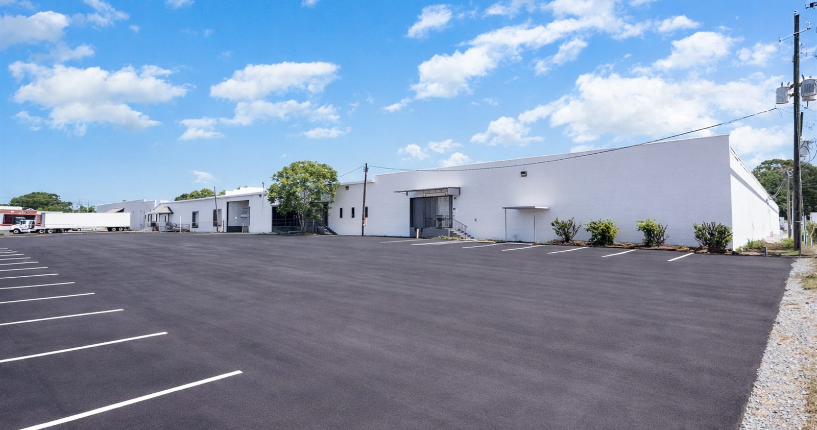 Ground-level view of the white-painted warehouse elevation at 2613 Lee Avenue across the newly paved and striped parking lot, with a tractor trailer parked at the far end