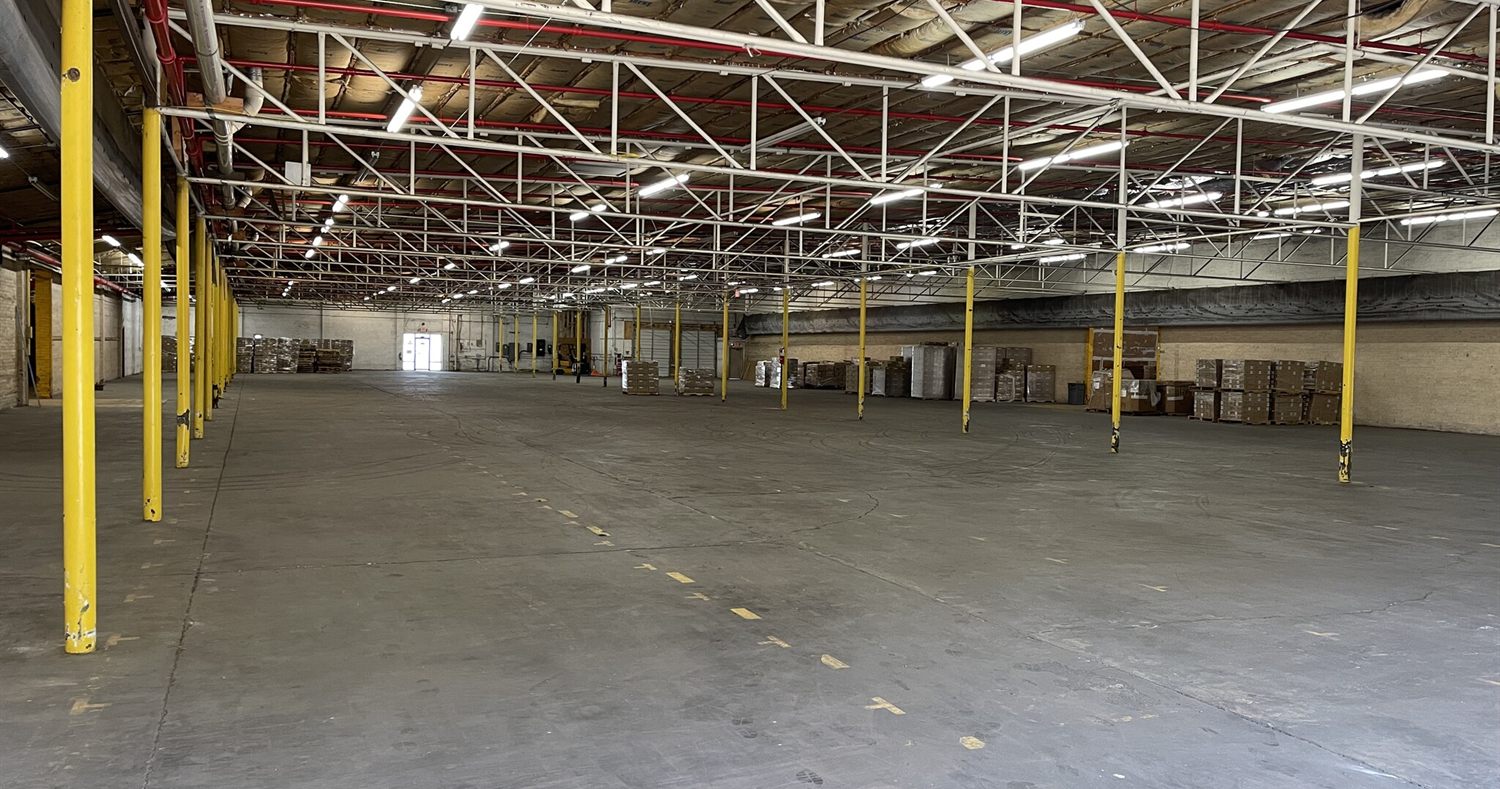 Wide clear-span bay at 2613 Lee Avenue under white bar-joist trusses and high-bay lighting, with yellow column guards and a striped concrete floor running toward the far wall
