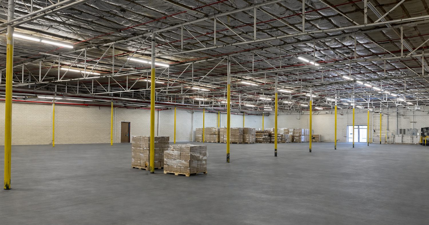 Open warehouse interior at 2613 Lee Avenue with painted brick walls, yellow column guards, a sealed concrete floor, and rows of fluorescent lighting under the insulated deck