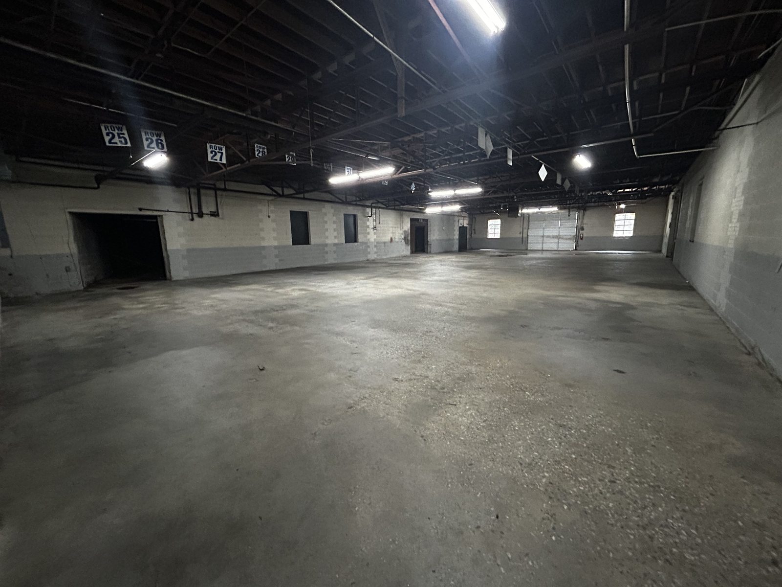 Interior of an available suite at 314 Mandustry Street: open warehouse with a concrete floor, painted block walls, exposed steel roof framing, and rows of fluorescent lighting