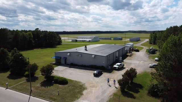 Drone view of 4475 Technology Drive NW, a gray metal building with a gravel drive along one side, and airport hangars and a runway behind it