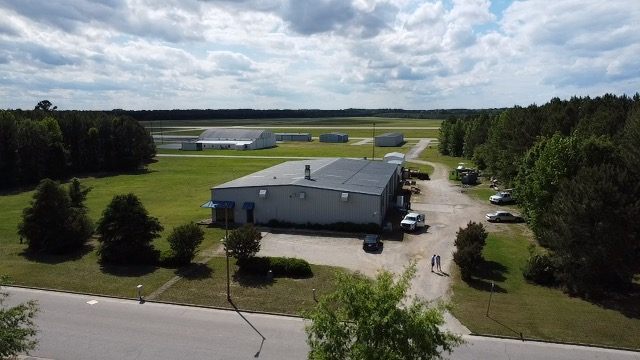Drone view of 4475 Technology Drive NW from the street, with the lawn, parking area, and gravel drive in front of the building and hangars in the distance