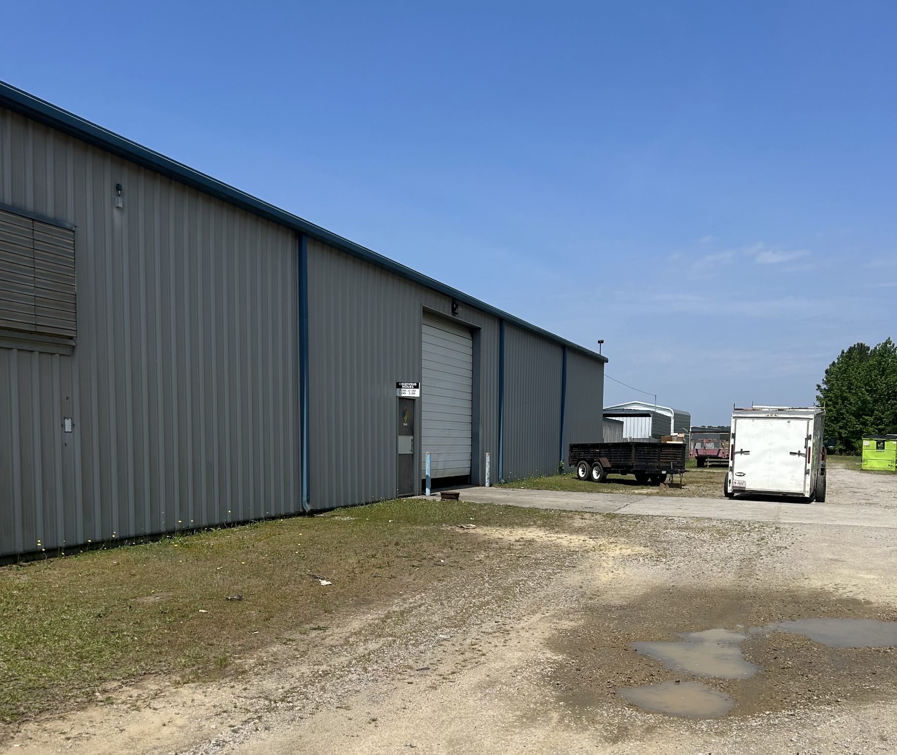 Long gray metal wall of 4475 Technology Drive NW with a white roll-up drive-in door, a gravel and concrete apron, and trailers parked beyond