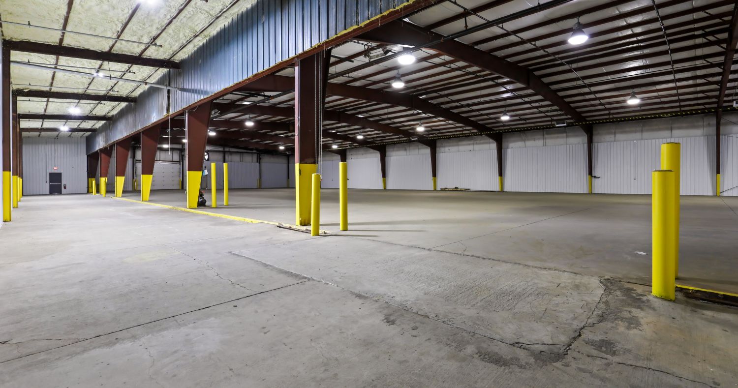 Clear-span warehouse interior at 6863 NC-56 with bowed steel trusses, high-bay lighting, painted column guards, and a sealed concrete floor