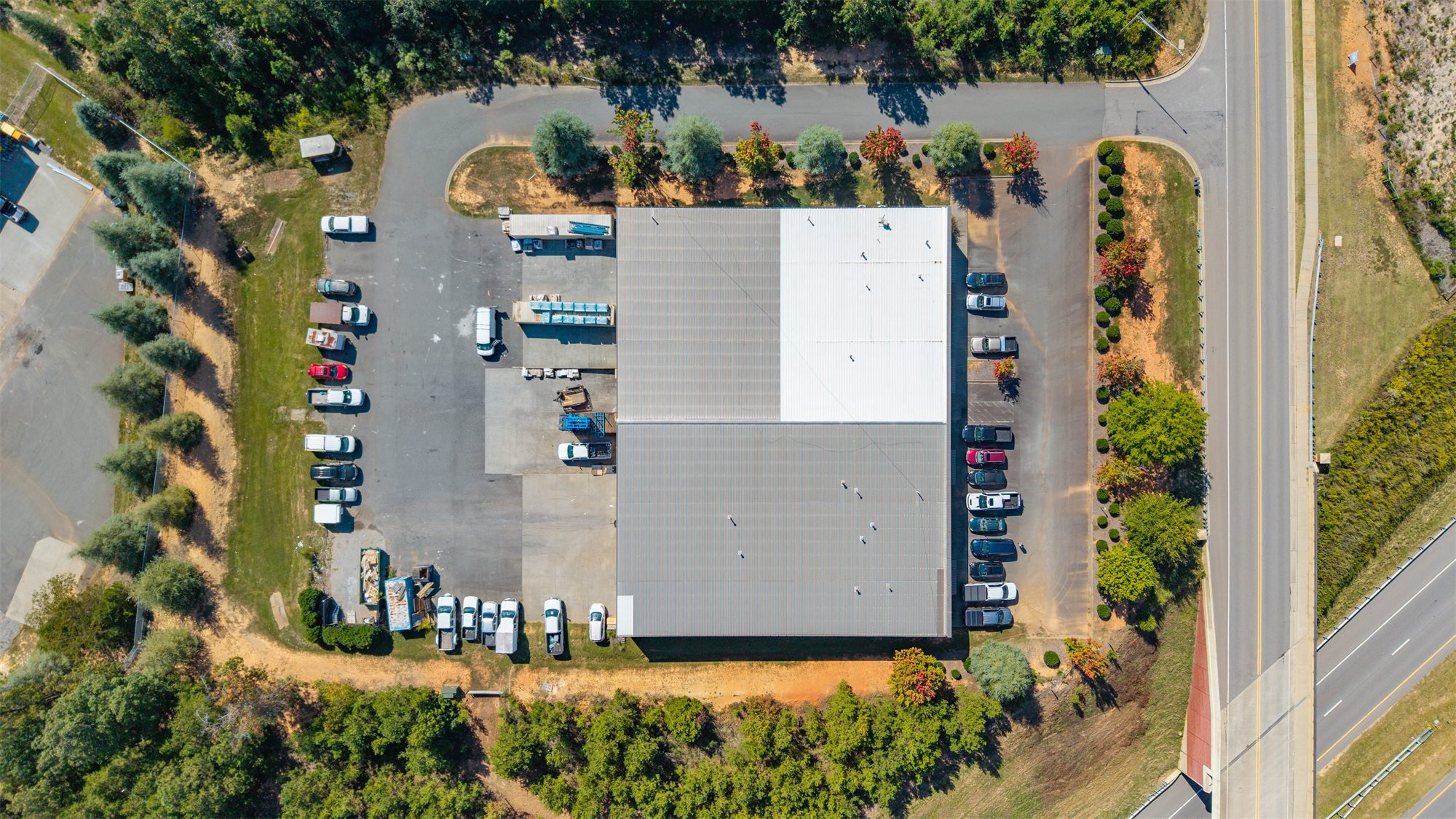 Overhead aerial of 7204 Stinson Hartis Road showing the rectangular metal-roofed building, a paved loading court and parking on one side, parking along the other, and Stinson Hartis Road rising onto the overpass beside it