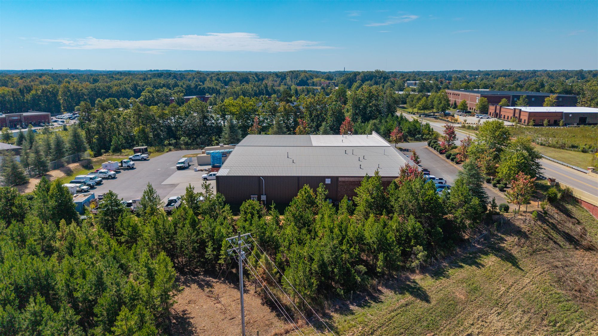 Low aerial of 7204 Stinson Hartis Road from the expressway side, with the metal warehouse behind a stand of pines and brick industrial buildings beyond