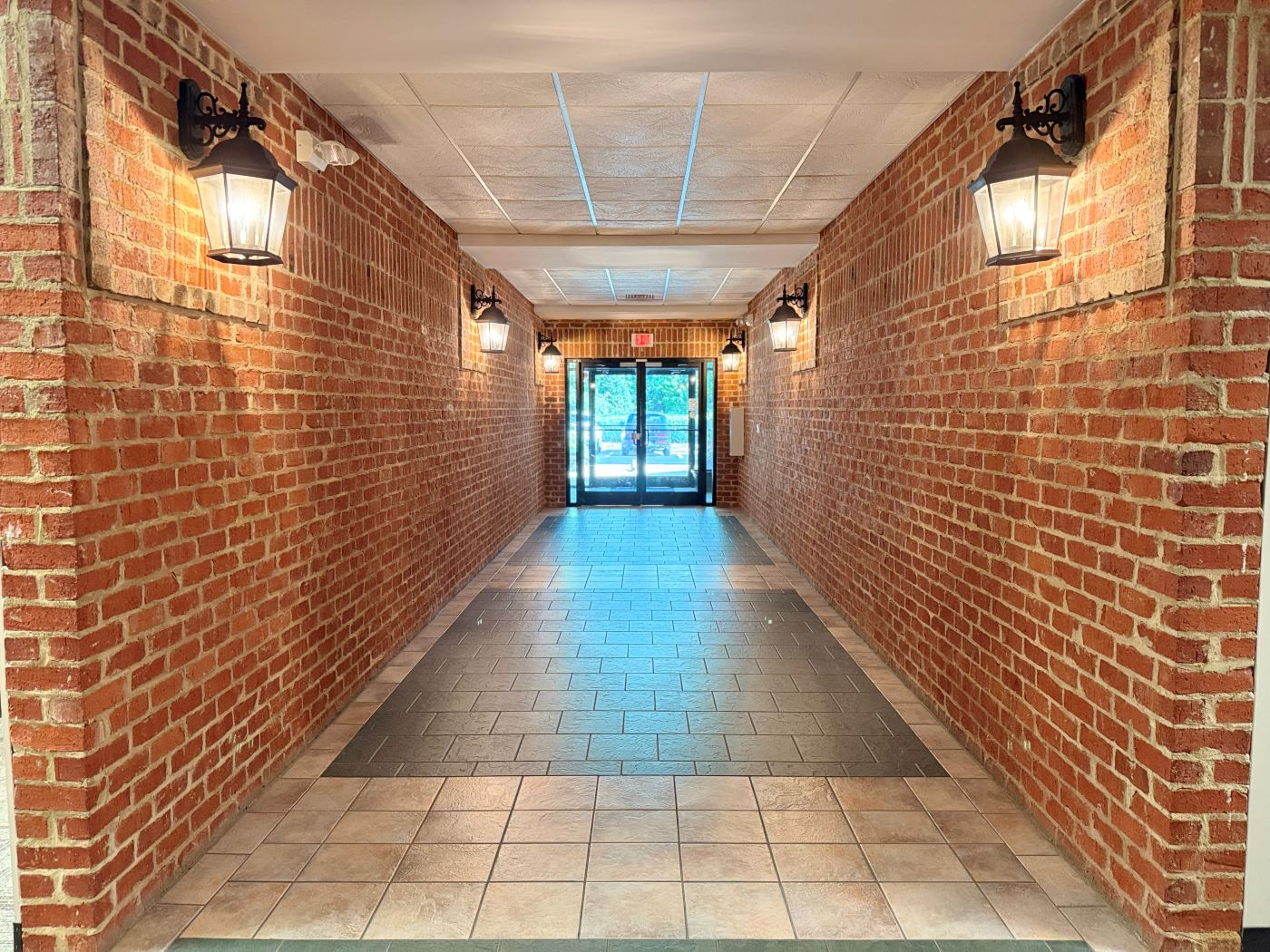 Covered brick walkway at Lochmere Pavilion with lantern sconces and tile flooring leading to the glass ground-floor entry of Suite 104