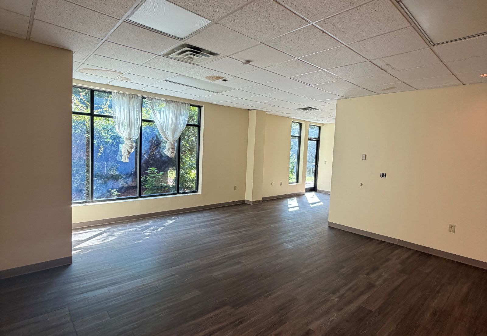 Open main room in Suite 104 at Lochmere Pavilion with new luxury vinyl tile flooring, freshly painted walls, and a wall of full-height windows looking onto the wooded rear of the site