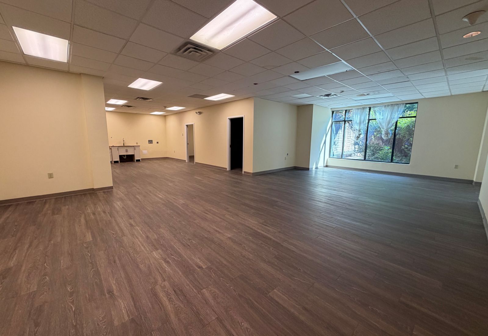 Suite 104 looking across the new luxury vinyl tile flooring and freshly painted walls toward the kitchenette, with doorways to the private rooms and a full-height window at right