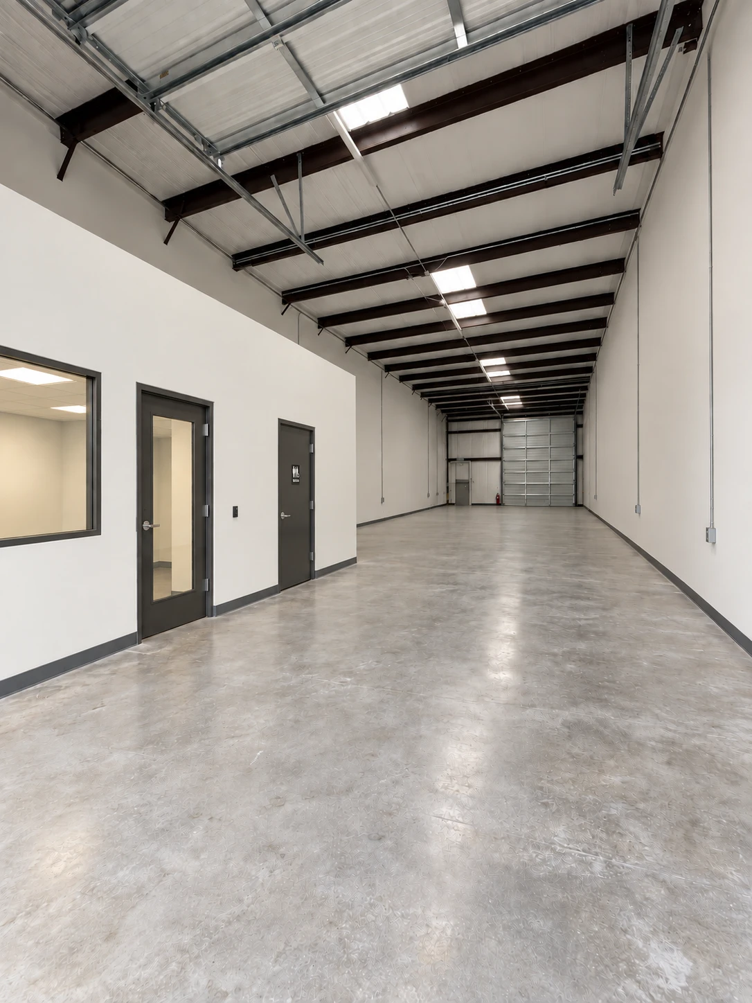 Interior rendering of a Building A suite: a long clear-span warehouse with polished concrete floors, exposed steel beams, a private office with a glass door and interior window, a restroom, and a drive-in overhead door at the far end
