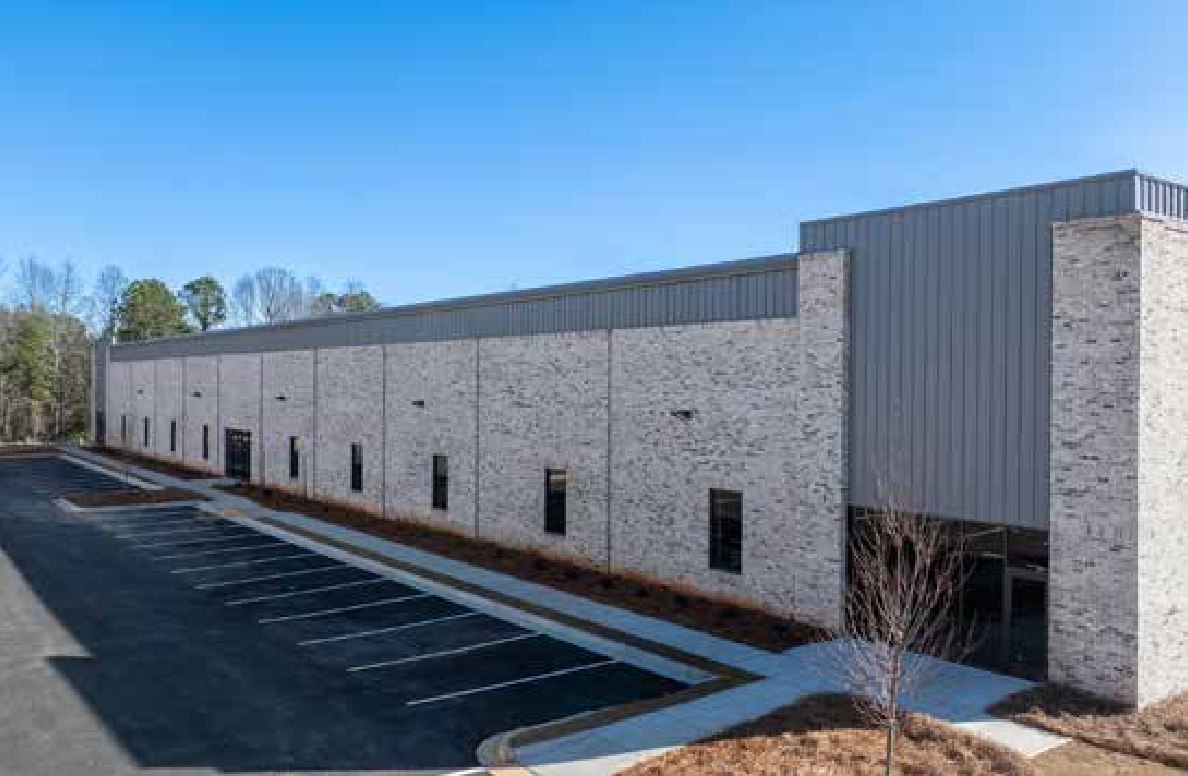 74 South Business Park, the completed Woodlands shallow-bay prototype, viewed along its length, showing light brick veneer, dark standing-seam metal, punched windows, and a striped parking field